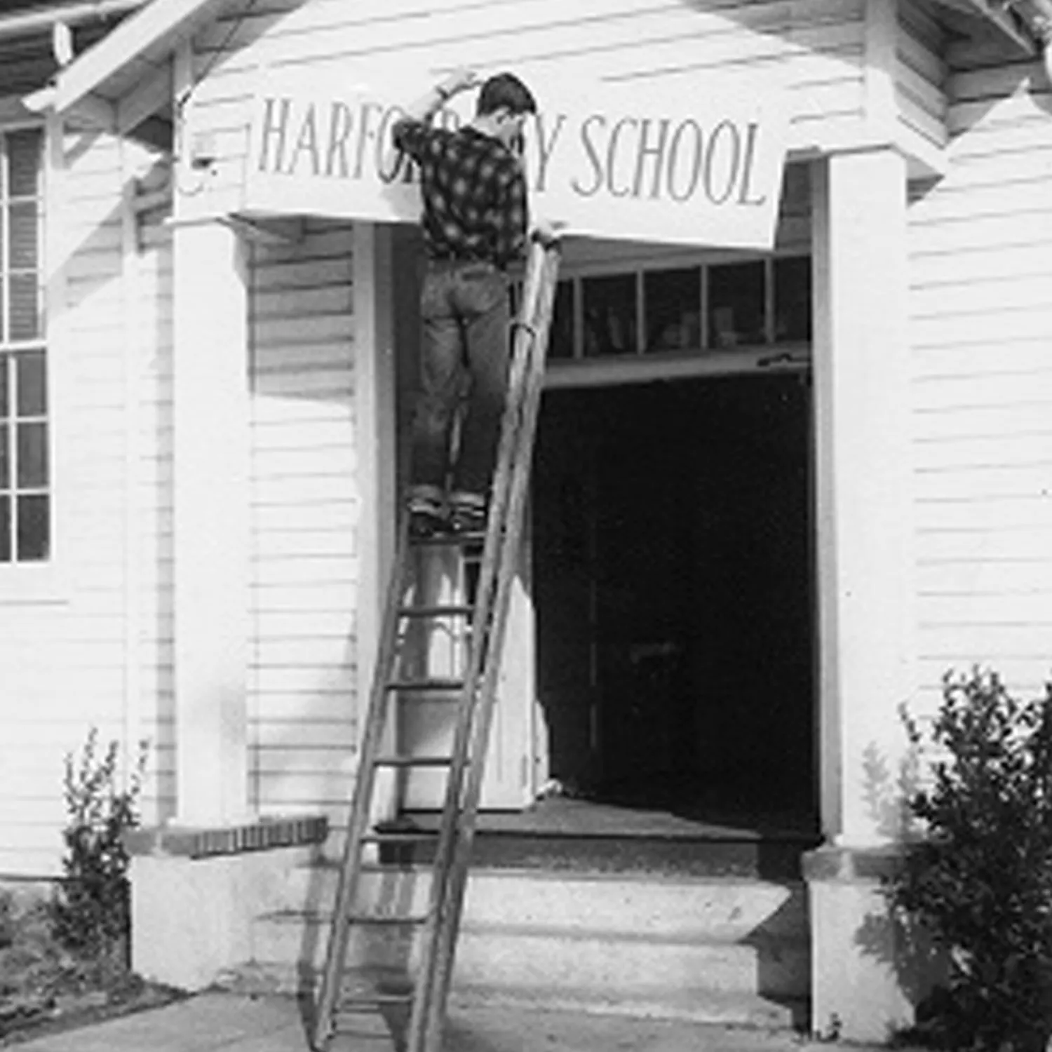 Black and white historical photo of a man hanging a sign for Harford Day School over the door of a schoolhouse